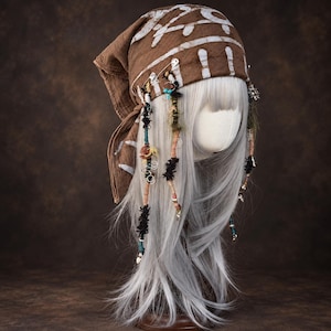 May include: A brown headscarf with white tribal-style patterns worn over a long, straight, silver-white wig. The headscarf is adorned with silver safety pins, small metal rings, turquoise beads, and dangling fabric strips in various colours.