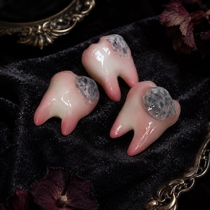 Puede incluir: Tres pequeños objetos brillantes con forma de diente hechos de un material rosa pálido. Cada diente presenta un área texturizada, de color gris oscuro y con cráteres en la superficie superior, que se asemeja a una caries.