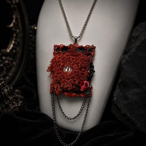 May include: A rectangular pendant necklace featuring a textured red organic surface with a central eye and a small red skull accent. The piece is framed by a black border and includes two draped silver-toned chains hanging from the bottom edge.