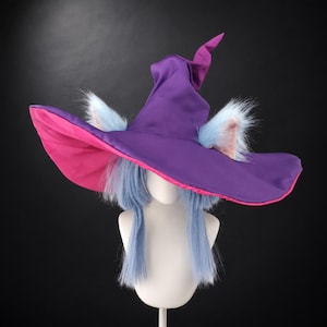 May include: A pointed witch hat featuring a deep purple exterior and a vibrant pink underside. The hat is adorned with two fluffy, light blue faux fur animal ears. Beneath the brim, long, straight, light blue synthetic hair hangs down.