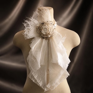 May include: An off-white fabric neck accessory featuring a layered, frayed-edge design. A central, rolled fabric rose with dark, distressed edges serves as the focal point, with additional textured, mesh-like fabric extending outwards.