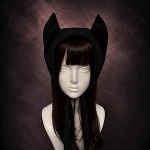May include: A black bonnet featuring two prominent, upright cat-like ears on the top. The headpiece is constructed from a solid black fabric and includes two long, matching black fabric ties that hang down from the chin area to secure the item.