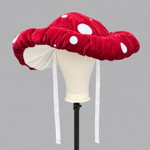 Puede incluir: Un sombrero de ala ancha con forma de seta, que presenta un exterior de terciopelo rojo intenso con puntos circulares blancos. La parte inferior del sombrero está forrada con tela blanca plisada e incluye dos cintas blancas largas que cuelgan.