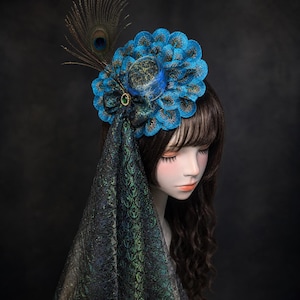 Op de afbeelding: Een fascinator met een cirkelvormige opstelling van blauwe, geschulpte stoffen bloemblaadjes met gouden accenten, een enkele pauwenveer, een kleine groene edelsteen en een lange, donkere, iriserende groen en zwart patroon stoffen sluier.