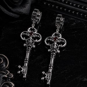May include: A pair of silver-toned metal earrings featuring a dangling key design. Each earring has a circular hoop top connected to an ornate, skeleton-style key pendant. A small, round, deep red gemstone is set in the center of each key head.