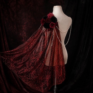 May include: A sheer black cape featuring a red spiderweb pattern, draped over a mannequin shoulder. The garment is adorned with a cluster of dark red and black artificial flowers and features delicate white pearl bead chains draped across the back.