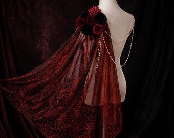Op de afbeelding: Een doorschijnende zwarte cape met een rood spinnenwebpatroon, gedrapeerd over de schouder van een etalagepop. De cape is versierd met een cluster van donkerrode en zwarte kunstbloemen en heeft fijne witte parelkettingen over de rug.