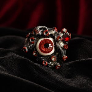 May include: A ring featuring a central white eyeball with a detailed red iris and black pupil, surrounded by dark, textured tentacles with small red circular accents and a single faceted red gemstone embedded on the right side of the band.