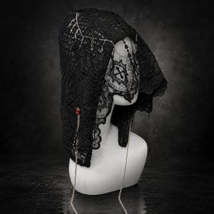 May include: A black lace head covering featuring an intricate floral pattern, a scalloped edge, and a veil section. The accessory is adorned with silver-toned chains, small cross charms, and a single circular red gemstone accent on the side.