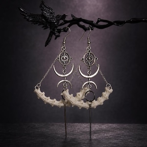 May include: A pair of silver-toned dangle earrings featuring a central eye charm, a crescent moon symbol, and a curved, off-white bone segment. The earrings are suspended by thin silver chains and include a straight metal bar hanging from the base.