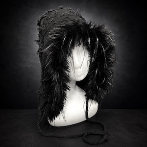 Könnte beinhalten: Eine schwarze Kapuze mit einer strukturierten, abgenutzten Stoffoberfläche und einem dicken Besatz aus langflorigem Kunstfell um die Gesichtsöffnung. Die Kapuze ist mit zwei langen, geflochtenen schwarzen Kordeln versehen, die herabhängen.