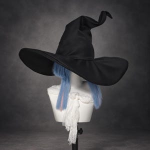 May include: A black witch hat with a wide brim and a pointed, slightly curved top. The hat is displayed on a white mannequin head featuring straight, light blue hair with a single pink streak, draped with a white lace fabric around the neck.