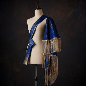 May include: A royal blue sash featuring a metallic gold speckled pattern throughout the fabric. The garment is draped over a mannequin and is finished with long, shimmering gold fringe trim that hangs from the lower edges of the blue fabric.