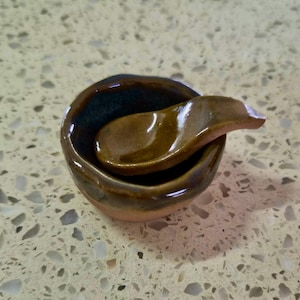 Op de afbeelding: Een kleine, ronde, handgemaakte keramische kom met een glanzend bruin en donkerblauw glazuur, met een bijpassende keramische lepel die in de kom rust. De buitenkant van de kom toont een natuurlijke, ongeglazuurde kleibasis met een onregelmatige rand.