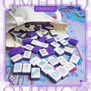 May include: A set of Mahjong tiles in a beige bag, scattered on a floral patterned surface. The tiles are white, purple, and green, with various symbols and characters. The bag has the text "OMHJGO".