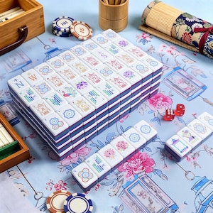 May include: A complete Mahjong set with white tiles featuring colorful floral and character designs. The tiles are stacked in a box with blue and purple stripes. Includes dice and game chips on a floral patterned surface.