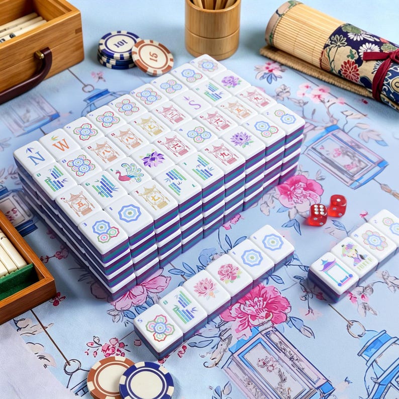 May include: A complete Mahjong set with white tiles featuring colorful floral and character designs. The tiles are stacked in a box with blue and purple stripes. Includes dice and game chips on a floral patterned surface.