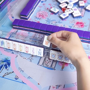 May include: A close-up of a Mahjong set with purple tiles and a floral patterned background. A hand is holding a tile with the letter "W" on it. The set includes tiles with various symbols and numbers.