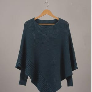 May include: A teal-colored knitted poncho with a round neckline and sleeves. The poncho is hanging on a wooden hanger against a neutral background. The knit pattern is visible, and the sleeves have a textured design.