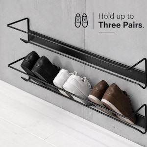 May include: A black metal wall-mounted shoe rack with two horizontal tiers. The rack holds three pairs of shoes: one pair of black dress shoes, one pair of white sneakers, and one pair of brown sneakers. Text on the wall reads: Hold up to Three Pairs.