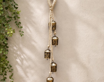 May include: A wall hanging decoration featuring five weathered, brass-colored metal bells suspended at varying heights from a thick, natural-colored twisted jute rope. The rope forms a loop at the top for hanging, with individual strands.
