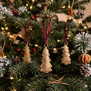 May include: Three metallic Christmas tree ornaments with a textured, aged gold finish. Each ornament is shaped like a stylized evergreen tree and hangs from a dark red fabric ribbon loop, suspended from the branches of a green pine tree.