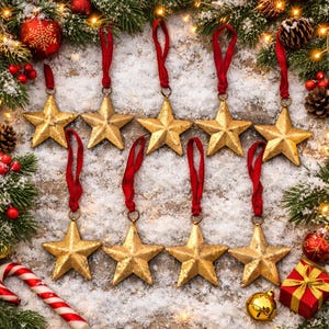 May include: A set of nine gold-colored star ornaments, each featuring a textured metallic finish and a red fabric ribbon loop for hanging. The stars are arranged in two rows against a snowy surface decorated with pine branches and holiday accents.