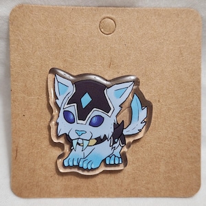 Puede incluir: Un pin de acrílico transparente con una ilustración estilizada de un felino dientes de sable azul claro con marcas de color púrpura oscuro, grandes ojos azul oscuro y un collar amarillo, sujeto a una tarjeta de cartón marrón cuadrada.