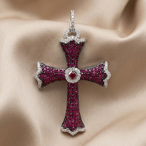 May include: A cross pendant featuring a dark metal base densely pavé-set with numerous small, vibrant red stones. The edges and the central circular motif are accented with clear, sparkling white stones, including a single square-cut red stone.