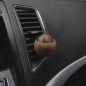 May include: A wooden car air freshener with a spherical design. The air freshener is brown and sits inside a small, matching wooden bowl, placed on a car's air vent. The car's interior is black, with a silver trim around the vent.