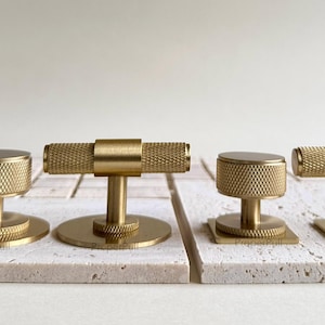 May include: Four brass cabinet knobs and pulls. Two knobs have a round base and a cylindrical top with a textured pattern. The other two have a T-bar design with a textured grip. All are mounted on off-white stone tiles.
