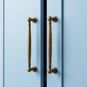 May include: Two vertical antique brass cabinet pulls with a smooth, elongated cylindrical center and rounded ends, mounted on light blue cabinet doors. The metal hardware features a weathered, dark gold finish with circular base plates.