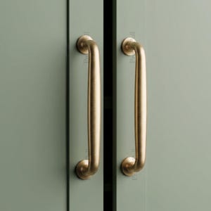 May include: Two vertical cabinet pulls with a brushed brass finish mounted on sage green cabinet doors. Each handle features a smooth, rounded bar design with circular mounting bases at the top and bottom, providing a clean and functional grip.