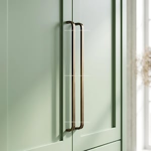 May include: Two long, vertical, bronze-toned metal cabinet pulls with a rectangular profile and rounded edges, mounted side-by-side on a light sage green cabinet door. The word ProperPull is faintly visible on the surface of the metal handles.