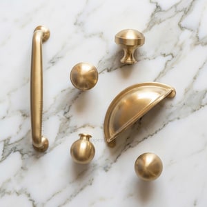 May include: A collection of brushed gold hardware including one long vertical handle, one semi-circular cup pull, and four spherical knobs of varying shapes. Each piece features a smooth, metallic finish and is arranged on a white surface.