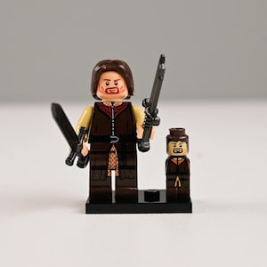 May include: Two Lego minifigures, one large and one small, of a warrior character. The larger figure holds two black swords and wears a brown tunic over a tan shirt. The smaller figure stands beside it, also in a brown tunic.