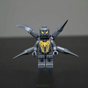 Op de afbeelding: Een grijze Lego-minifiguur met een spinnenthema. De figuur heeft een geel spinnenembleem op de borst en gele ogen. Het heeft beweegbare armen en vier extra beweegbare aanhangsels die zich van de rug uitstrekken. Het woord "Minifigures" staat onderaan gedrukt.
