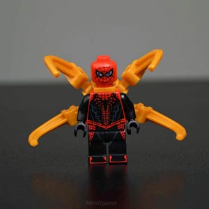 Può includere: Minifigure Lego di Spider-Man in tuta nera e rossa con ali meccaniche dorate. La figura ha una testa rossa con un disegno a ragnatela nera e occhi neri. La figura è su una superficie scura.