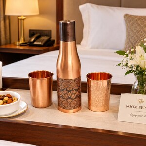 Peut inclure: Une bouteille d'eau en cuivre avec un bouchon en métal foncé et une section centrale à motifs noirs, entourée de deux gobelets en cuivre sur un plateau en bois. Le plateau contient un bol blanc de noix et une carte avec le texte ROOM SERVICE Enjoy your stay.