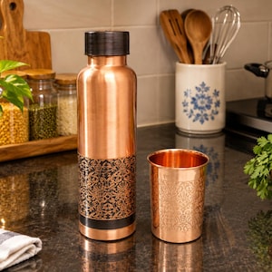 Peut inclure: Une bouteille d'eau cylindrique en cuivre avec un bouchon marron foncé et une bande noire ornée d'un motif floral complexe sur la partie inférieure, accompagnée d'un gobelet en cuivre assorti avec un motif floral en relief sur sa surface.