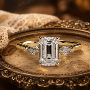 May include: A gold ring featuring a large emerald-cut centre stone held by four prongs, flanked by two smaller kite-shaped side stones. The band is polished yellow gold, and the setting is designed to showcase the clear, faceted gemstones.