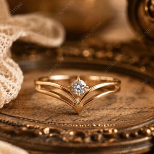 Op de afbeelding: Een gouden ring met een V-vormige band met een gespleten ontwerp en een enkele ronde, briljant geslepen heldere edelsteen die in het middelpunt is gezet. Het metaal heeft een gepolijste afwerking en de band loopt taps toe naar de achterkant.