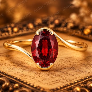Könnte beinhalten: Ein Goldring mit einem großen, oval geschliffenen, tiefroten Edelstein in einer Vier-Krappen-Fassung. Die Ringschiene hat eine glatte, polierte Oberfläche und ein geschwungenes Design, das den zentralen Stein umschließt und elegant wirkt.