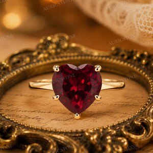 May include: A gold ring with a heart-shaped, deep red gemstone. The ring is set in a four-prong setting, and the band is a simple, polished gold. The ring is displayed on a decorative, ornate gold frame.