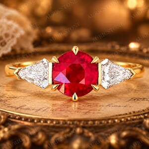 May include: A gold ring featuring a large, round, vibrant red centre stone held by six prongs, flanked by two clear, triangular-cut side stones. The band is polished yellow gold, creating a classic three-stone design with a bright, faceted appearance.