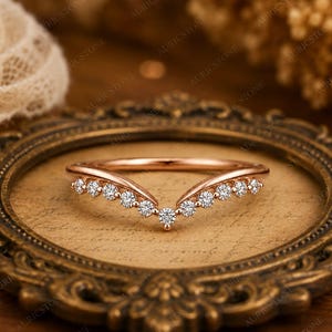 May include: A V-shaped rose gold ring featuring a series of small, clear, round-cut stones set along the chevron-style band. The metal has a polished finish, and the stones are arranged symmetrically, meeting at a central point in the middle.