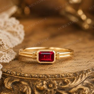 Könnte beinhalten: Ein Goldring mit einem rechteckigen, im Smaragdschliff geschliffenen roten Edelstein in einer Zargenfassung. Die Ringschiene hat eine glatte, polierte Oberfläche mit doppelten Linien auf beiden Seiten des zentralen Steins.