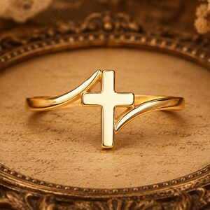 May include: A polished gold ring featuring a central cross design. The band consists of two smooth, curved metal segments that meet at the cross, creating a continuous, flowing shape. The surface of the gold is reflective and smooth throughout.