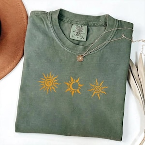 May include: An olive green short-sleeve t-shirt featuring a graphic of three yellow celestial symbols, including two suns and a crescent moon. The inner neck label displays the text Comfort Colors with size S P CH, and a gold-toned necklace rests on top.