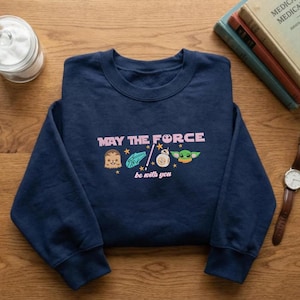 May include: A navy blue crewneck sweatshirt featuring the pink text "MAY THE FORCE be with you" and small graphic icons of Chewbacca, the Millennium Falcon, a lightsaber, BB-8, and Grogu, accented with several small yellow stars across the chest.